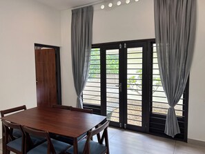 Une pièce avec une table à manger en bois, des chaises et une vue sur l’extérieur, visible à travers une porte munie de rideaux.