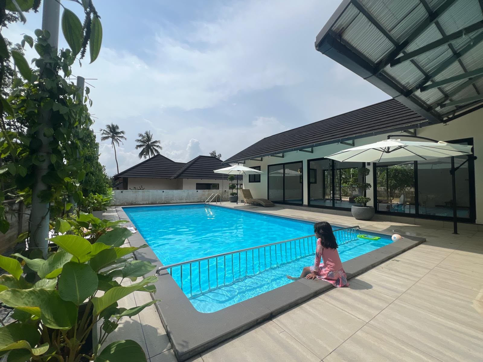 Piscina cercada por vegetação e arquitetura moderna, com uma criança sentada na borda.