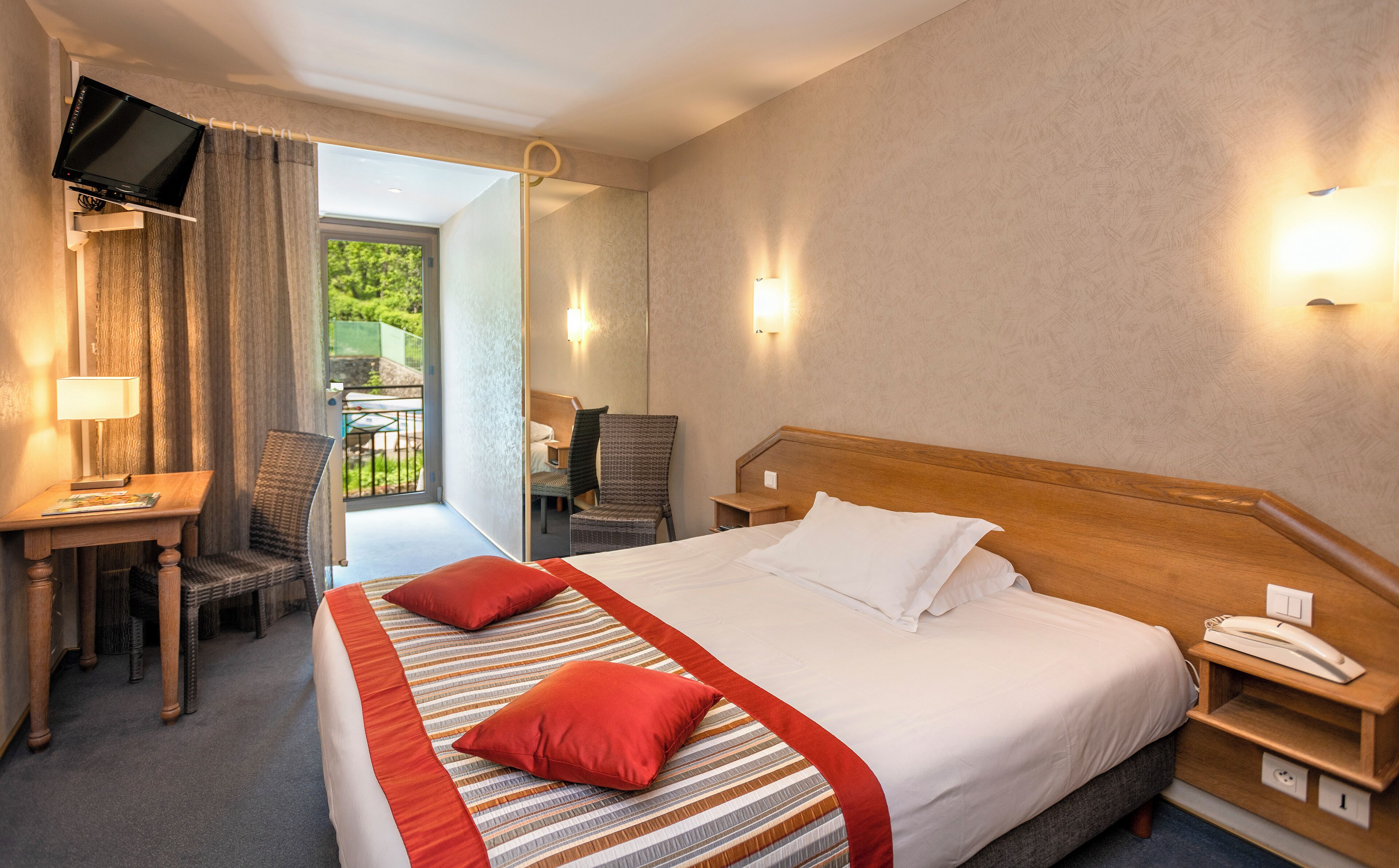 A hotel room with a bed, a desk, a chair, a TV, and a balcony with a view of greenery.