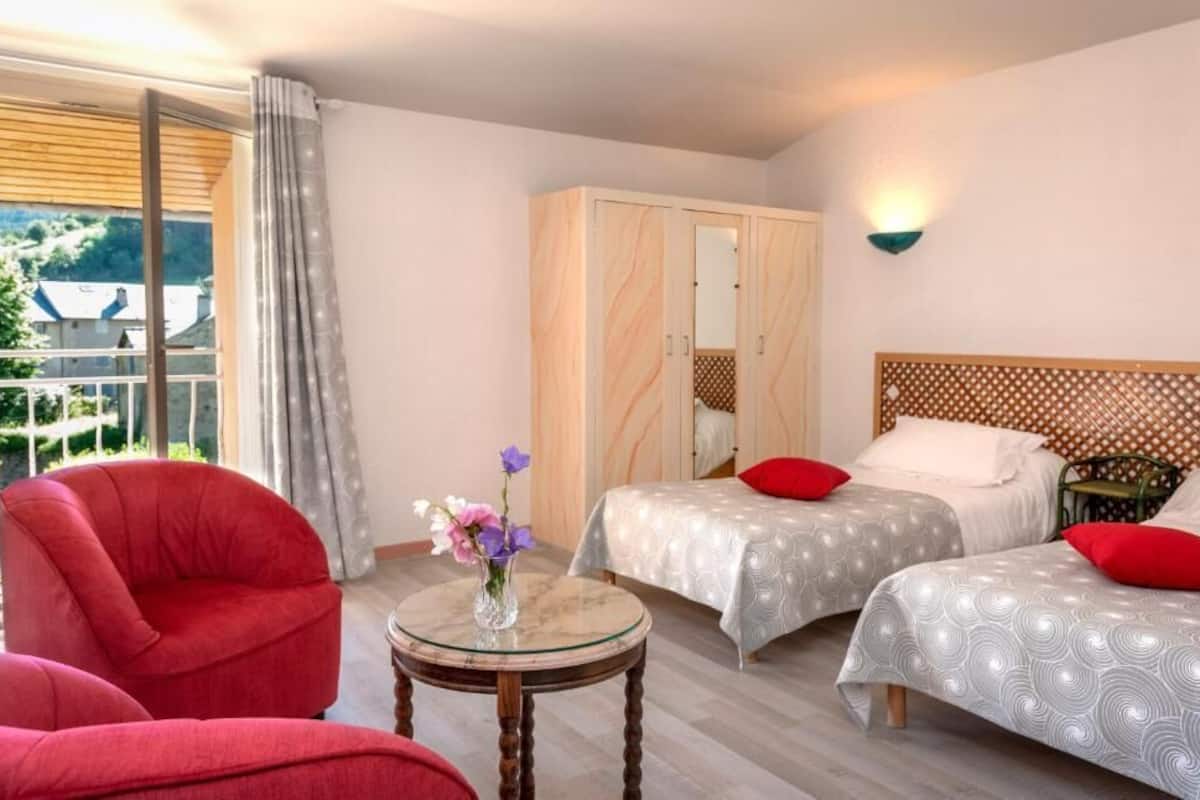 Une chambre d’hôtel avec deux lits, une chaise rouge, une petite table avec un vase, et une vue sur la verdure à l’extérieur.