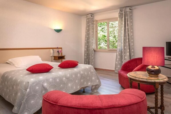 A hotel room with a bed, red chairs, a small table with a lamp, a window with curtains, and a wall-mounted light fixture. - Logis Hotel Restaurant Mont Aigoual (Meyrueis)