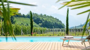 Ein Pool mit Holzterrasse, einer Liege und Blick auf eine üppige grüne Landschaft mit Bäumen und Hügeln.