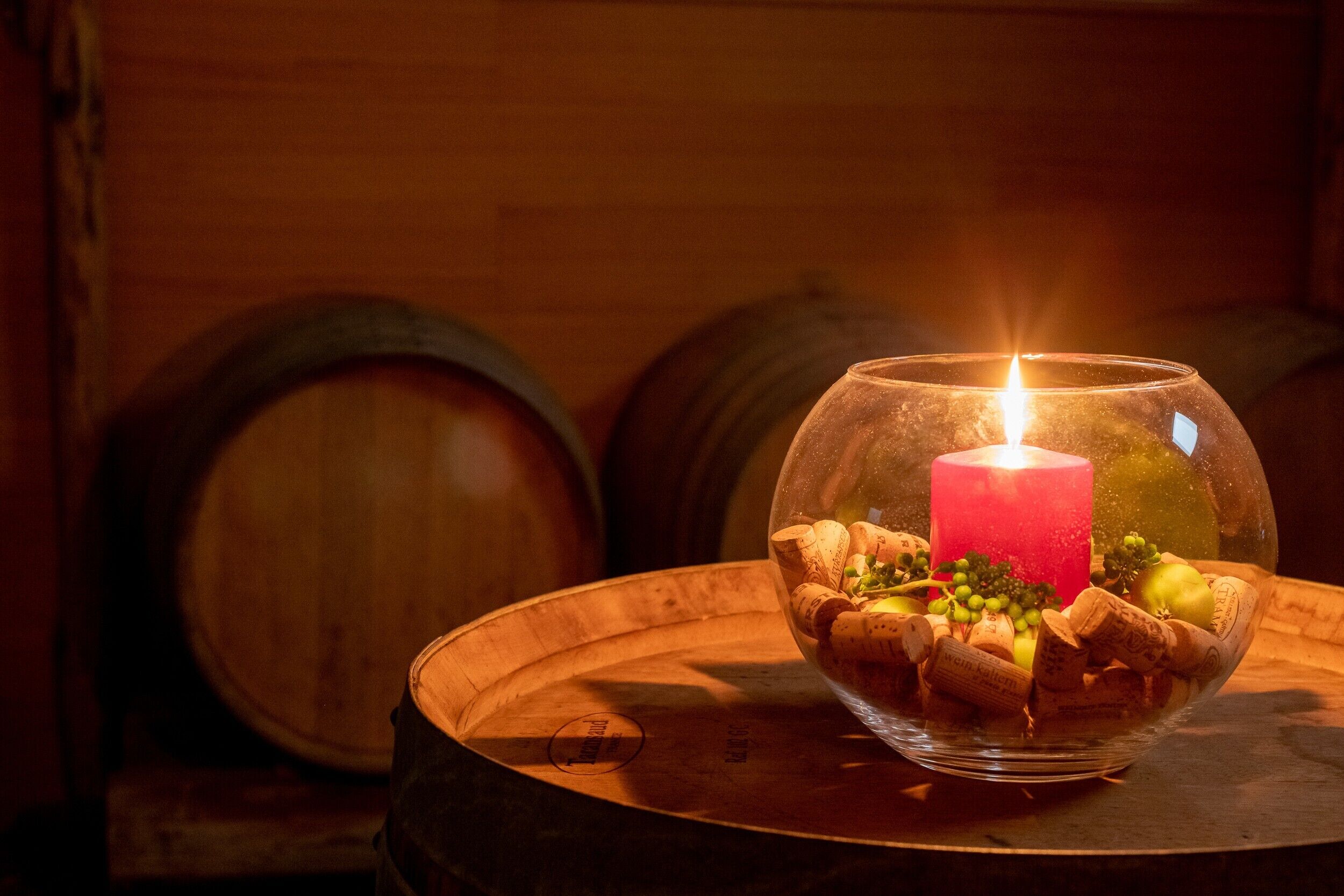 Une bougie dans un bol en verre, posée sur une table en bois, entourée de bouchons de bouteilles de vin et de quelques éléments de verdure.