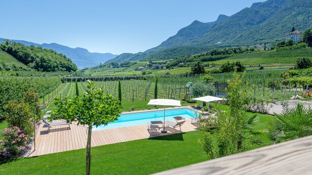 A pool with a wooden deck, surrounded by lush greenery and mountains in the background. - Apartment Kardonighof Mareit (Kaltern)