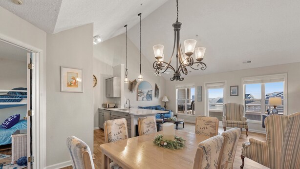 A spacious dining area with a wooden table and upholstered chairs, a chandelier, and a view of the kitchen and living room through an open doorway. - Buccaneer Village 734: Community Pool | Walking Distance to Marina | Building El (Manteo)