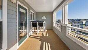 A covered balcony with a view of neighboring houses, a small table with chairs, and a door with a glass panel. - Buccaneer Village 734: Community Pool | Walking Distance to Marina | Building El (Manteo)