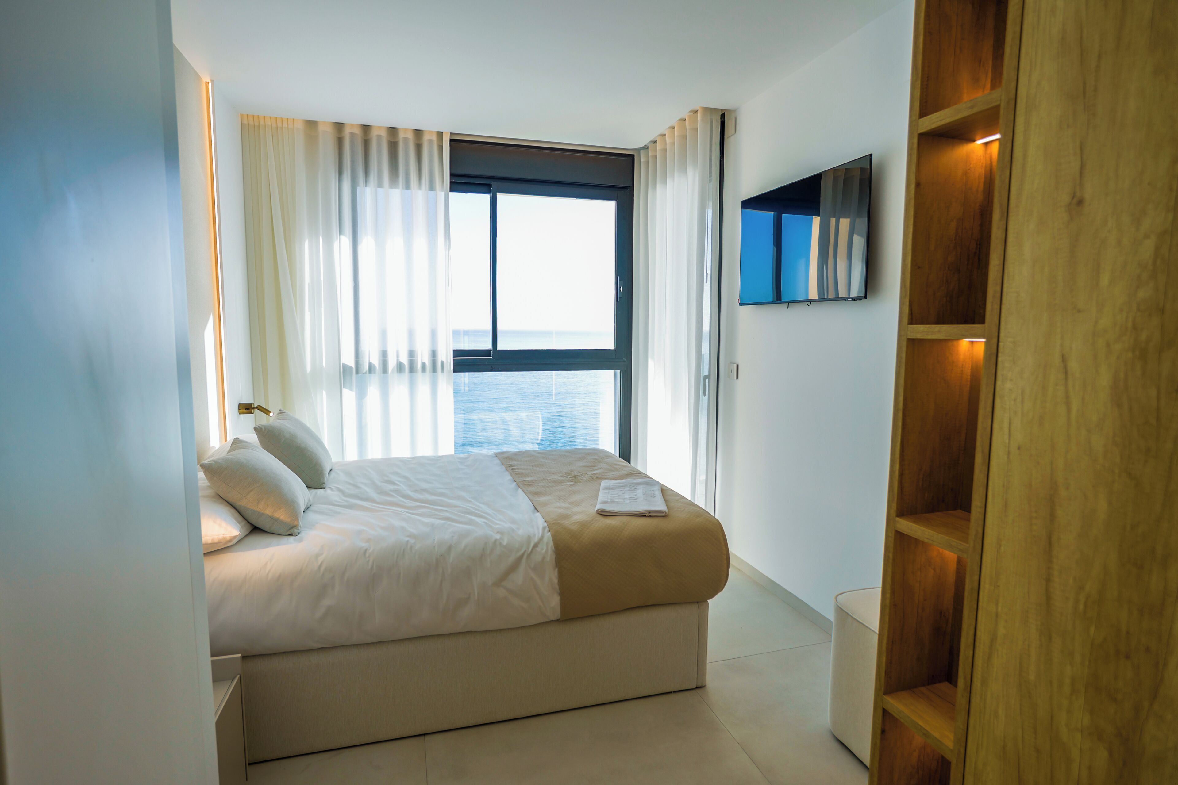 A modern bedroom with a large bed, a TV mounted on the wall, and a view of the sea through the window.
