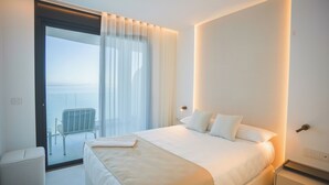 A modern hotel room with a large bed, a small bedside table, and a view of the sea through a sliding glass door.