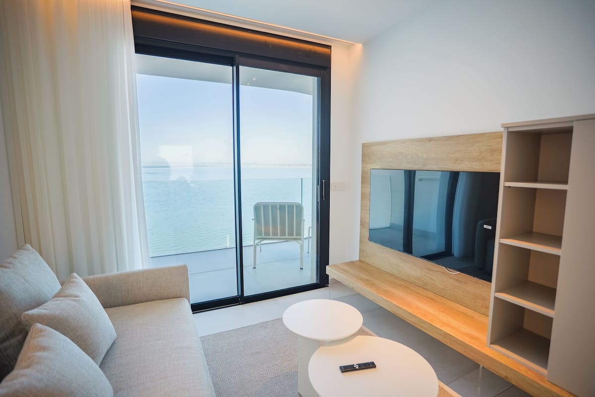 Un salon moderne avec un canapé, une table basse ronde, un téléviseur posé sur une étagère en bois et une porte-fenêtre coulissante donnant sur un balcon équipé d’une chaise.