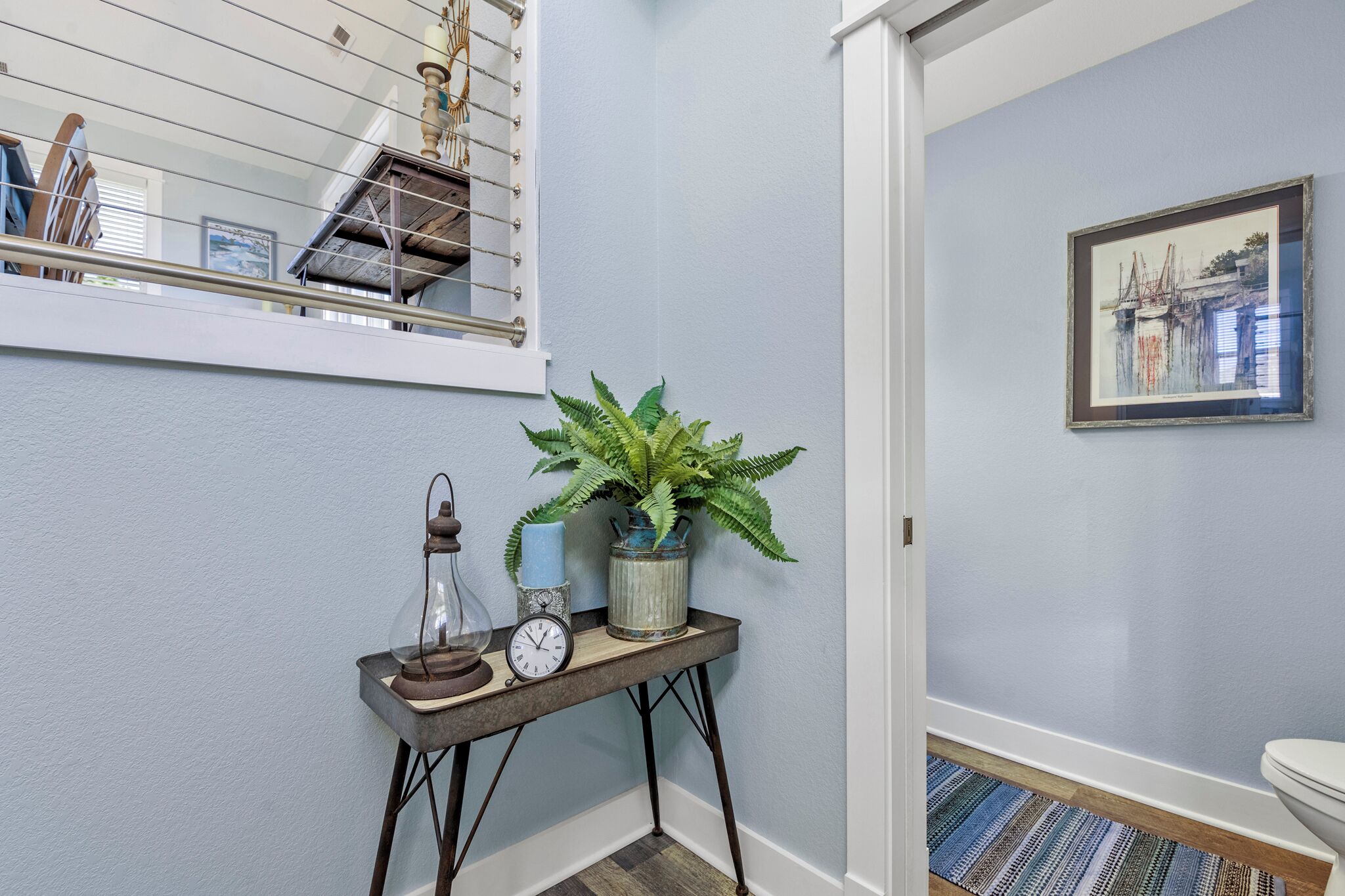 A hallway with a light blue wall, a framed picture, a potted plant, a small table with a clock, and a window with blinds.