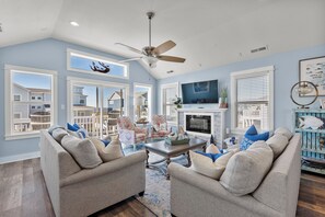 A spacious living room with a ceiling fan, large windows, a fireplace, and a television. - Sound Legacy: Private Pool and Hot Tub (Corolla)