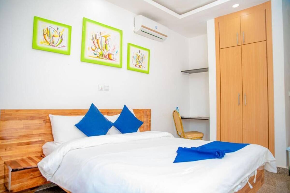 A bedroom with a wooden headboard, a bed with white bedding and blue pillows, a wooden wardrobe, and three framed artworks on the wall.