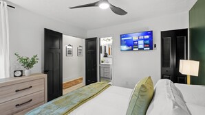Hotellihuone, jossa on sänky, kattotuuletin, seinälle asennettu televisio, yöpöytälamppu ja pieni kasvi lipaston päällä.