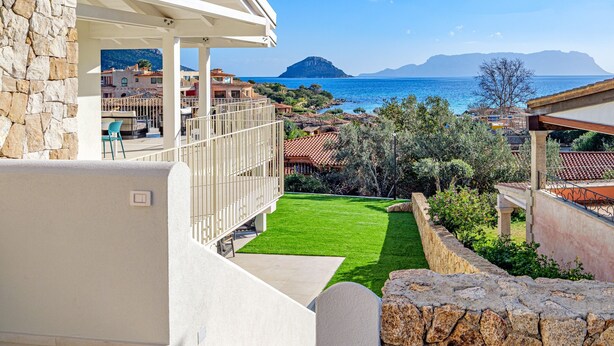 A balcony with a stone wall, a green lawn, and a view of the sea and mountains.