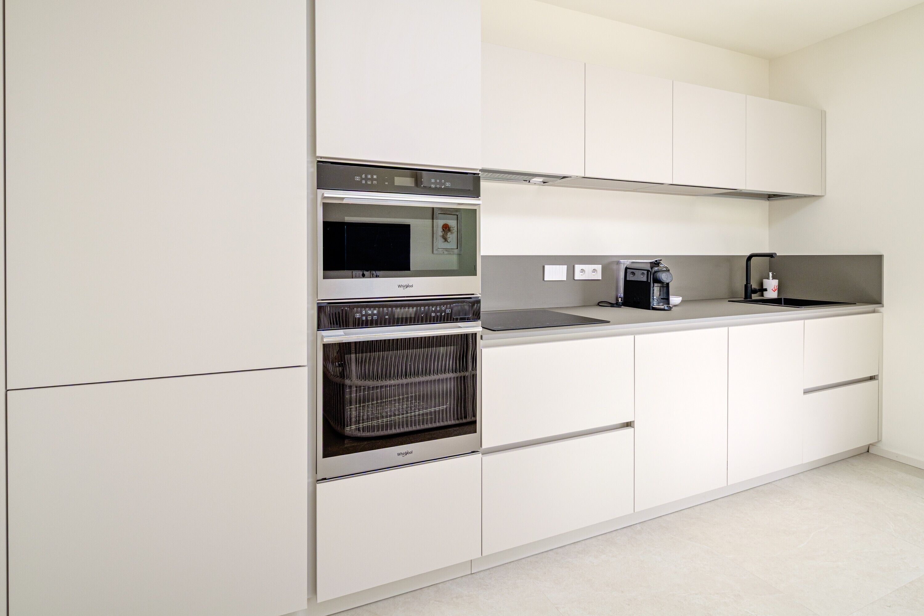 Una cocina moderna con muebles blancos, horno empotrado, cafetera y lavabo.