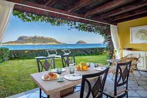 An outdoor dining area with a view of the sea and hills, featuring a table set with croissants, fruit, and a pitcher. - Blue Apartment - S' Abba e Sa Pedra with Sea View, Wi-Fi and Air Conditioning (Golfo Aranci)