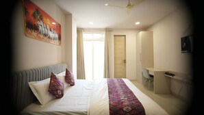 A hotel room with a bed, a desk, a chair, a ceiling fan, and a painting on the wall. - Hotel Joystel Studio (Jaipur)
