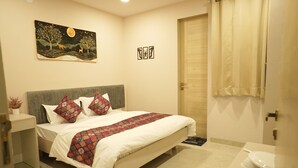A bedroom with a bed, a nightstand, a bedside table, a painting, a door, and a bathroom area. - Hotel Joystel Studio (Jaipur)
