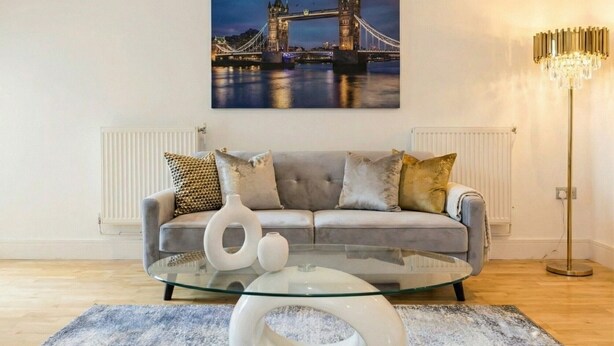 Uma sala de estar com um sofá cinza, uma mesa de centro de vidro, um vaso branco e um quadro de uma ponte.