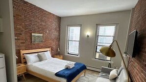 Ein Schlafzimmer mit einem hölzernen Bett, einem grauen Sofa, einem blauen Hocker und einer Ziegelwand.