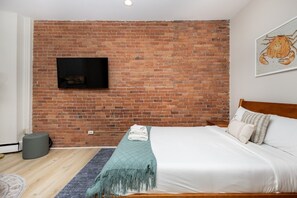 A bedroom with a brick wall, a flat-screen TV mounted above the bed, a wooden headboard, and a bed with white bedding and multiple pillows. - Peaceful Back Bay Studio (Boston)