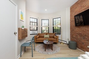A compact living space with a brown leather sofa, a round coffee table, a blue chair, and a small rug. There are three windows with blinds, a mounted TV, and a wooden cabinet with a plant. - Peaceful Back Bay Studio (Boston)