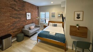 Quarto moderno com parede de tijolos, cama de madeira, sofá cinza e uma pequena escrivaninha.