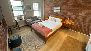 Um quarto com parede de tijolos, cama com manta vermelha, poltrona cinza, mesa de cabeceira de madeira e uma pequena mesa com abajur.