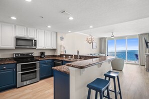 A modern kitchen with white cabinets, a granite countertop, stainless steel appliances, and a large island with bar stools. - Palazzo 1203 - Two Bedroom with bunk on the gulf (Panama City Beach)