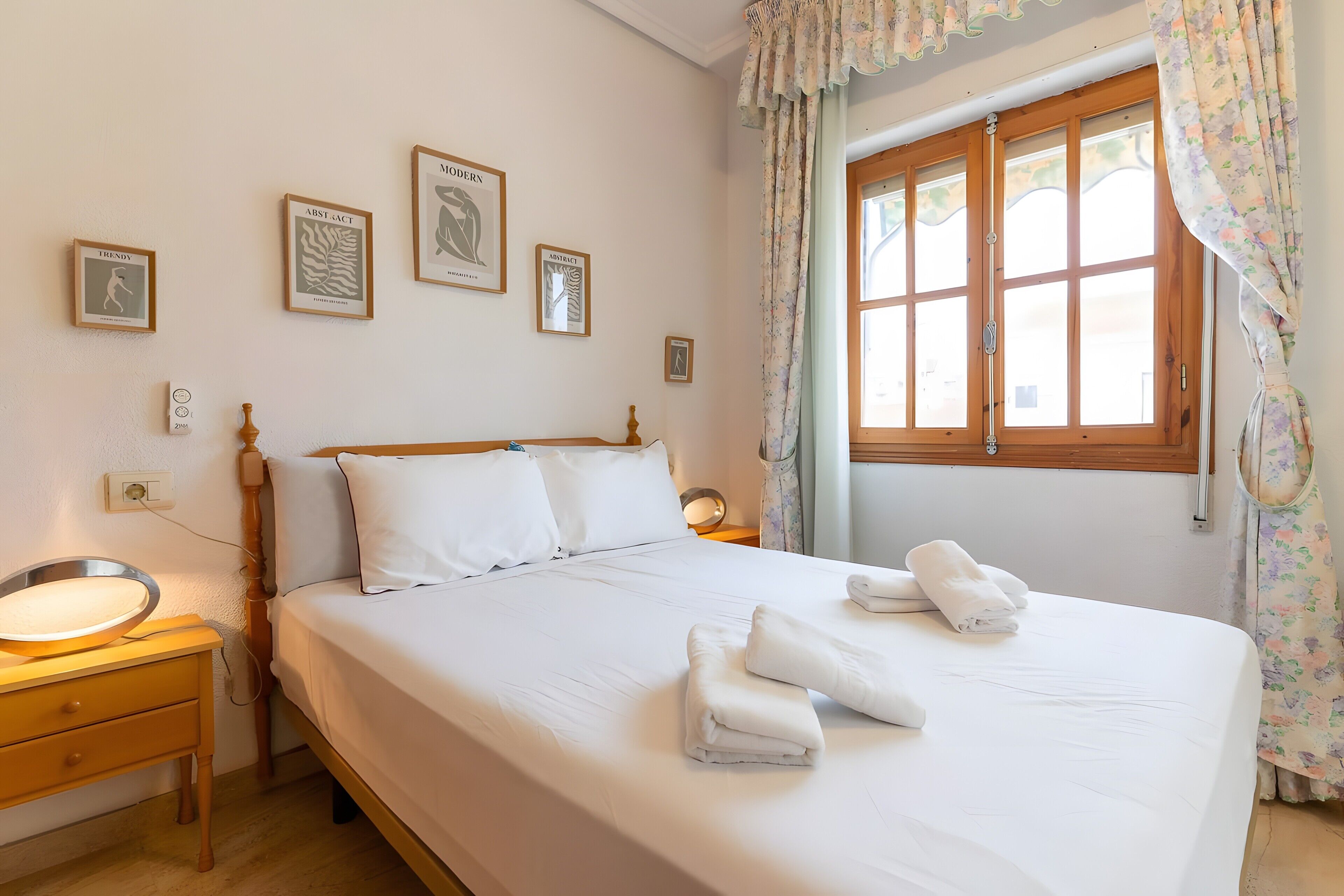 A neatly made bed with white linens and towels, a wooden headboard, and a window with floral curtains.