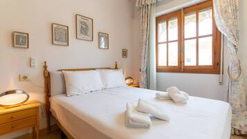 Un lit bien fait, avec du linge de lit et des serviettes blanches, une tête de lit en bois et une fenêtre ornée de rideaux à motifs floraux.