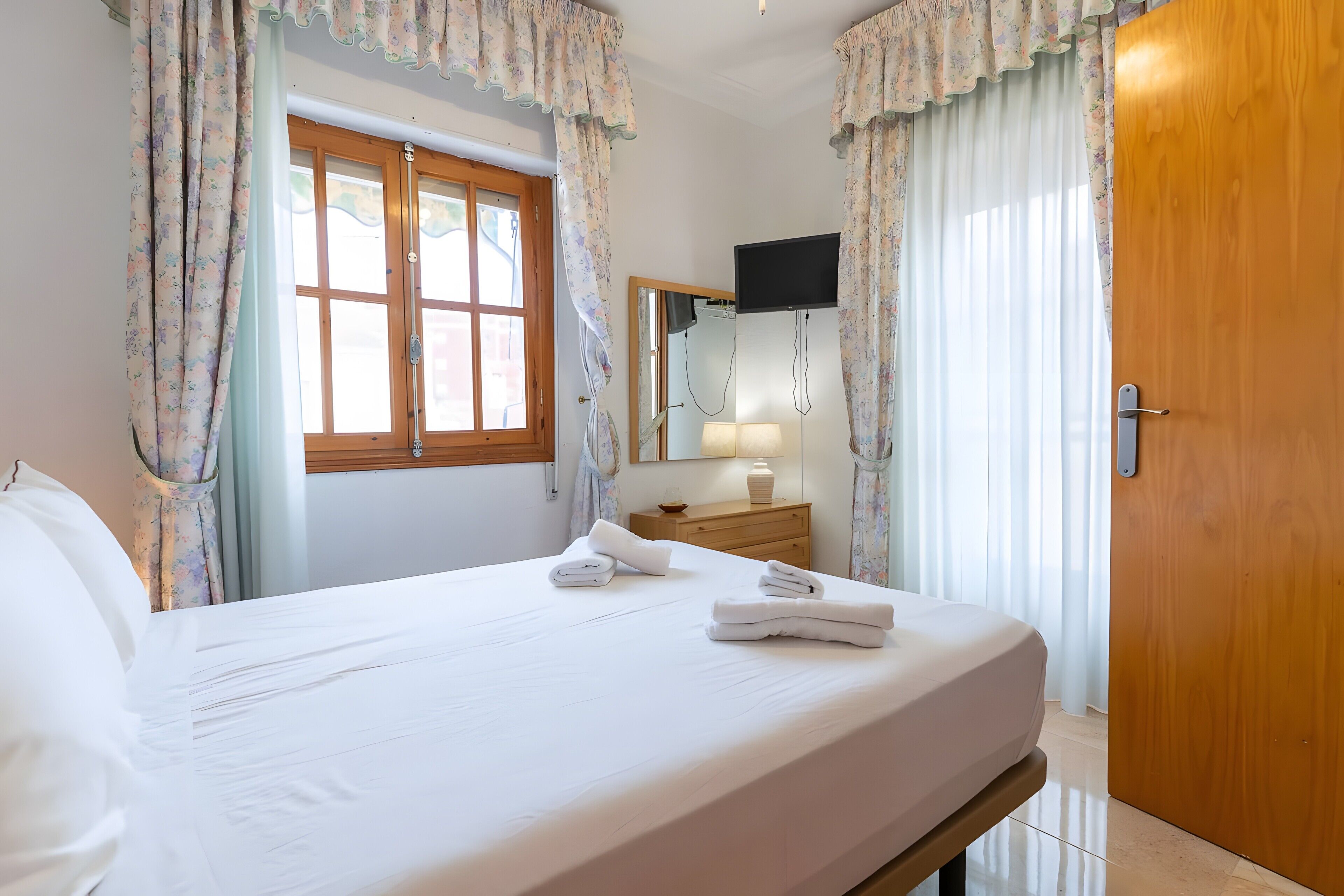 A neatly made bed with towels, a wooden door, a window with curtains, a bedside table with a lamp, and a TV mounted on the wall.