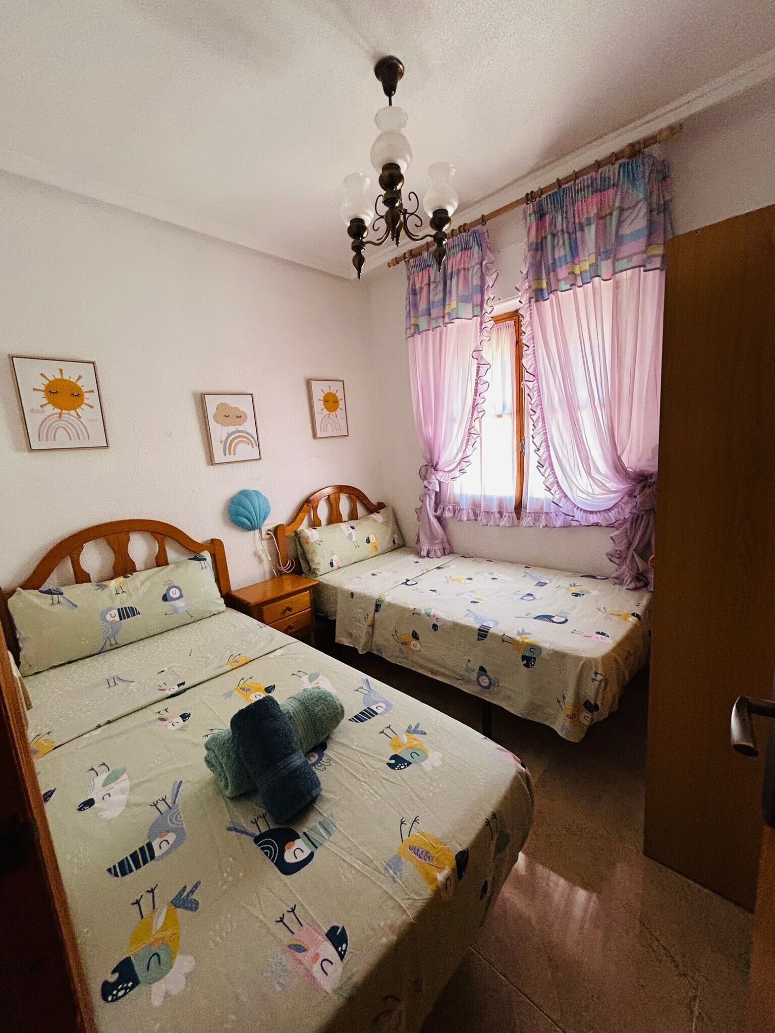 A bedroom with two beds, a chandelier, and a window with curtains.