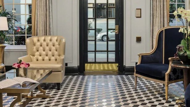 Een kamer met een zwart-wit schakeerd patroon op de vloer, een groot raam met gordijnen, een deur met een ruit en twee fauteuils.