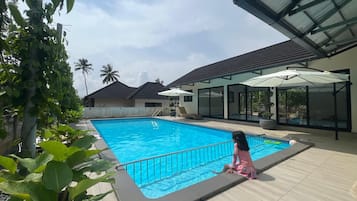 A swimming pool with a child sitting by the edge, surrounded by greenery and modern architecture.