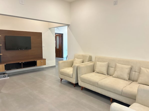 A modern living room with a large flat-screen TV mounted on a wooden wall, a beige sofa with multiple cushions, and a minimalist design.