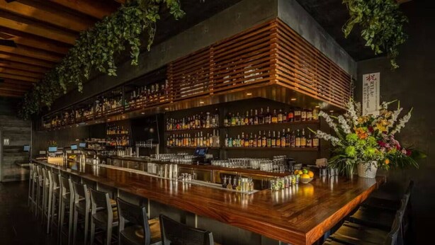 A bar with a wooden counter, metal shelves with bottles, and chairs. - Check in with a Cocktail | Heart of Hermosa Beach (Hermosa Beach)