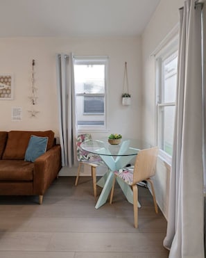 Ein gemütliches Wohnzimmer mit einem braunen Sofa, einem Esstisch mit Glasplatte und Stühlen sowie einem Fenster mit Vorhängen.