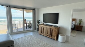A spacious living room with a large TV, a wooden cabinet, and a view of the beach. - Charming beachfront condo, Daytona Beach Shores (Daytona Beach Shores)
