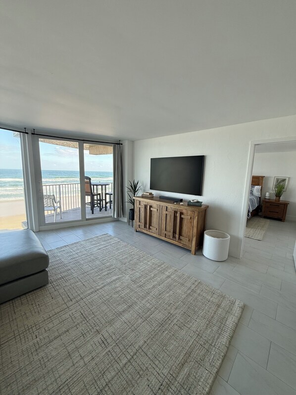 A spacious living room with a large TV, a wooden cabinet, and a view of the beach. - Charming beachfront condo, Daytona Beach Shores (Daytona Beach Shores)