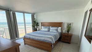 A bedroom with a large bed, a wooden desk, a nightstand, and a view of the beach. - Charming beachfront condo, Daytona Beach Shores (Daytona Beach Shores)
