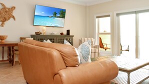 Un salon avec un canapé en cuir, un téléviseur fixé au mur, une table en bois et une vue sur la plage grâce à de grandes fenêtres.