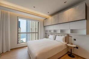 A modern hotel room with a large bed, a bedside table, and a view of tall buildings. - Murjan 2702 (Dubai)