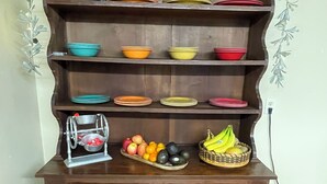 Un meuble en bois doté de plusieurs étagères, dont certaines contiennent des bols et des assiettes aux couleurs vives, et un panier rempli de fruits placé sur l’étagère inférieure.