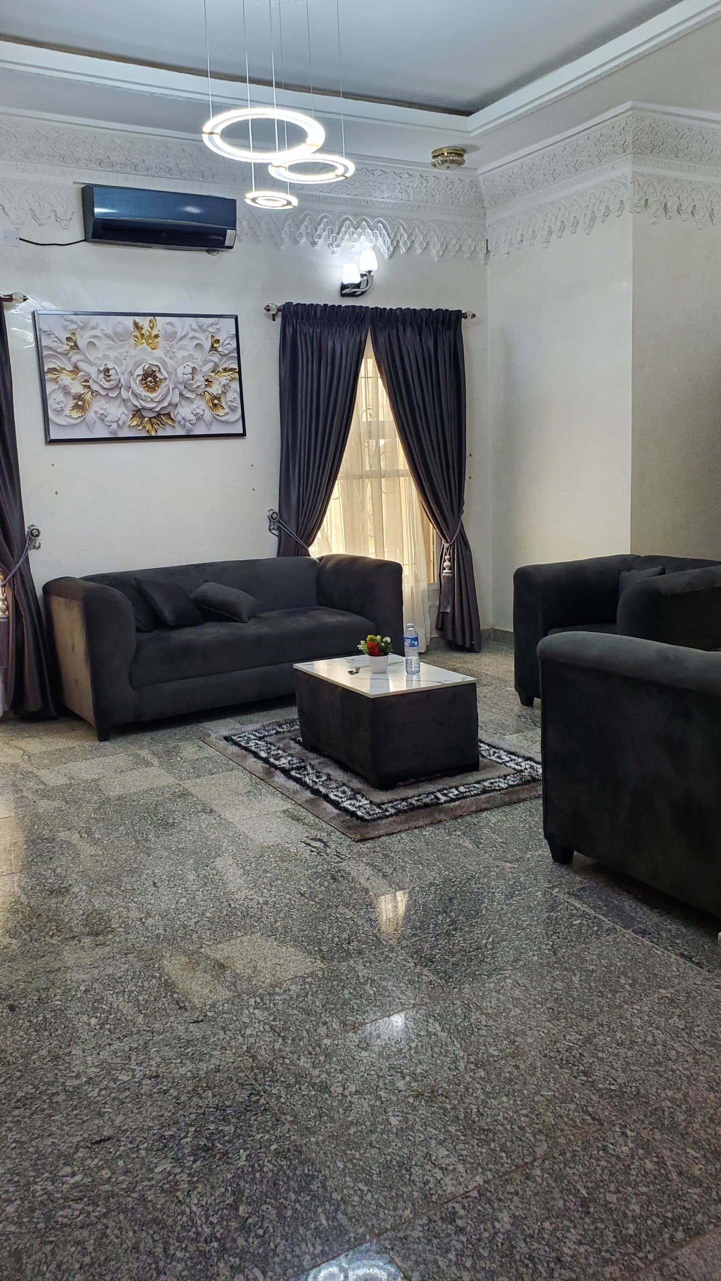A living room with a dark sofa, a coffee table, and a rug. There is a window with curtains, a wall-mounted air conditioner, and a decorative piece above the sofa.