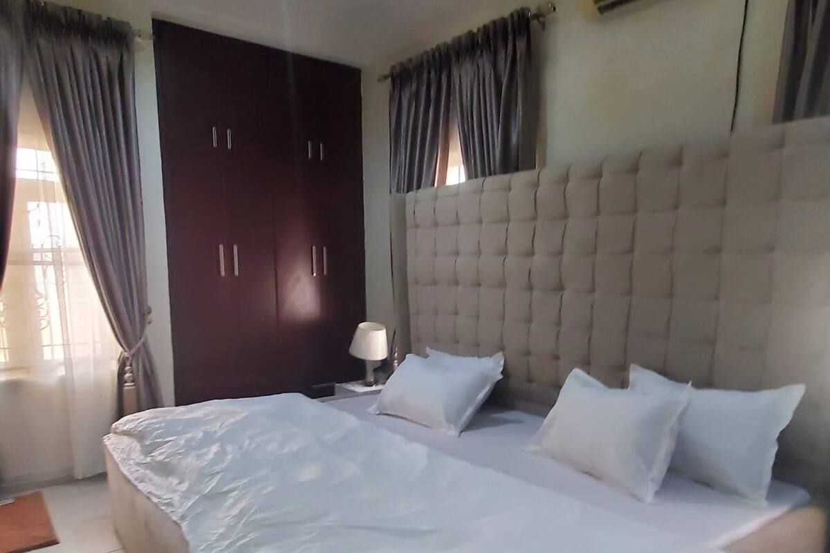 A bedroom with a large bed, a wardrobe, a bedside table with a lamp, and an air conditioning unit on the wall.