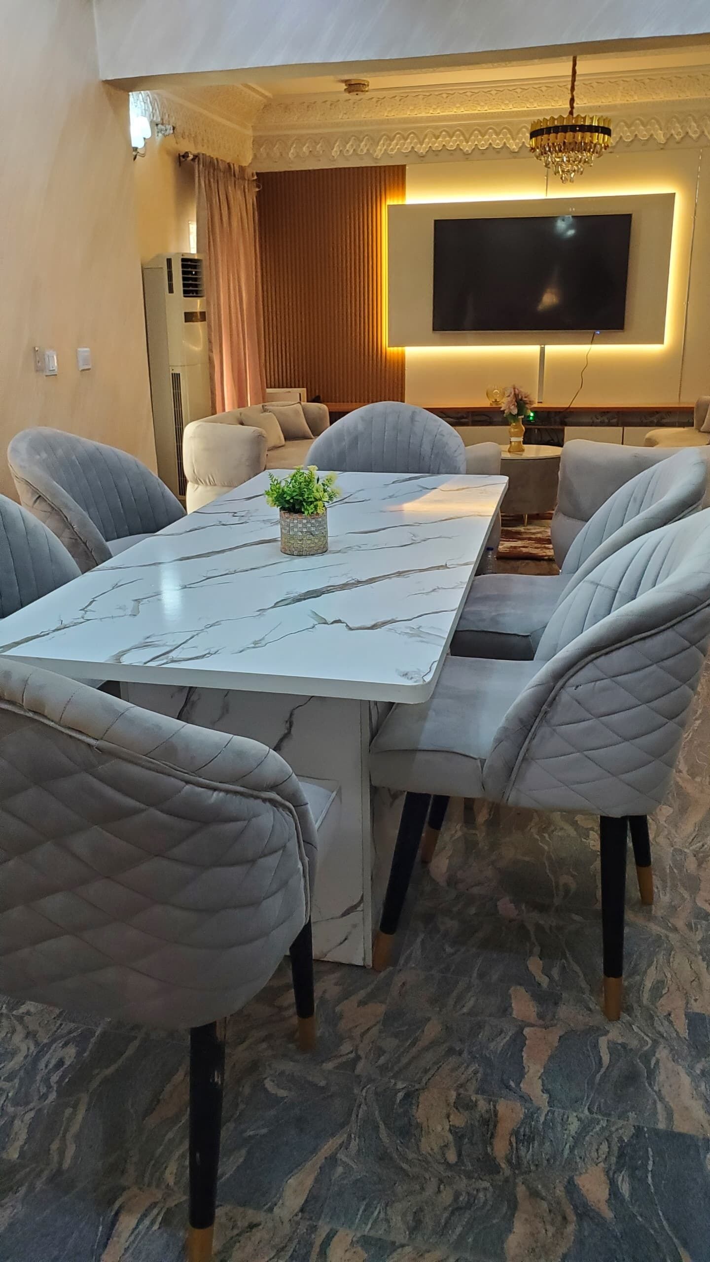 Un espace repas comprenant une table en marbre, des chaises rembourrées, un lustre et un téléviseur à écran plat.