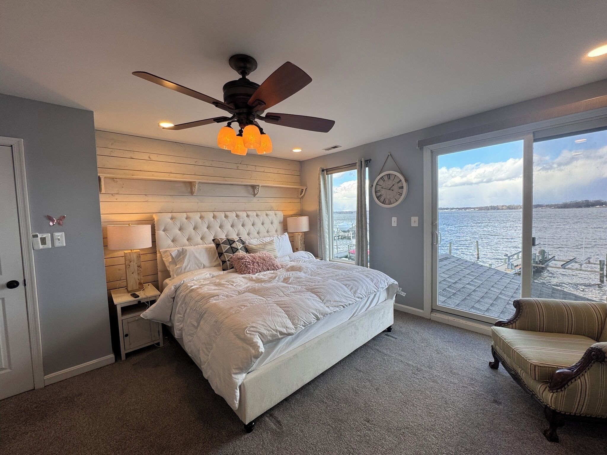 A bedroom with a large bed, a ceiling fan, a view of the water, and a clock on the wall.