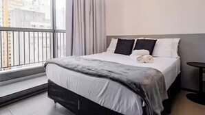 Ein modernes Hotelzimmer mit einem großen Bett, Blick auf die Stadt vom Balkon und einem kleinen Tisch am Fenster.