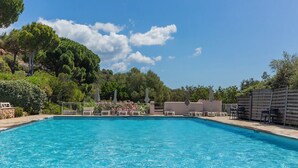 Una piscina rettangolare con lettini e ombrelloni sul bordo, circondata da vegetazione e un cielo sereno.
